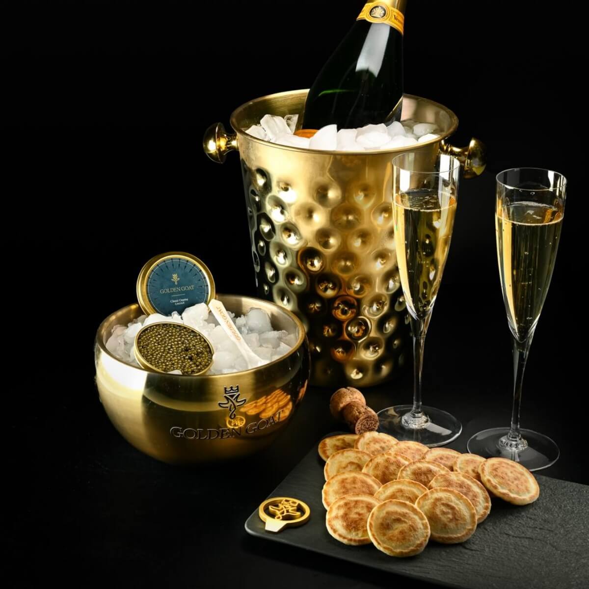 Gold champagne bucket with bottle, glasses, and caviar on a black background
