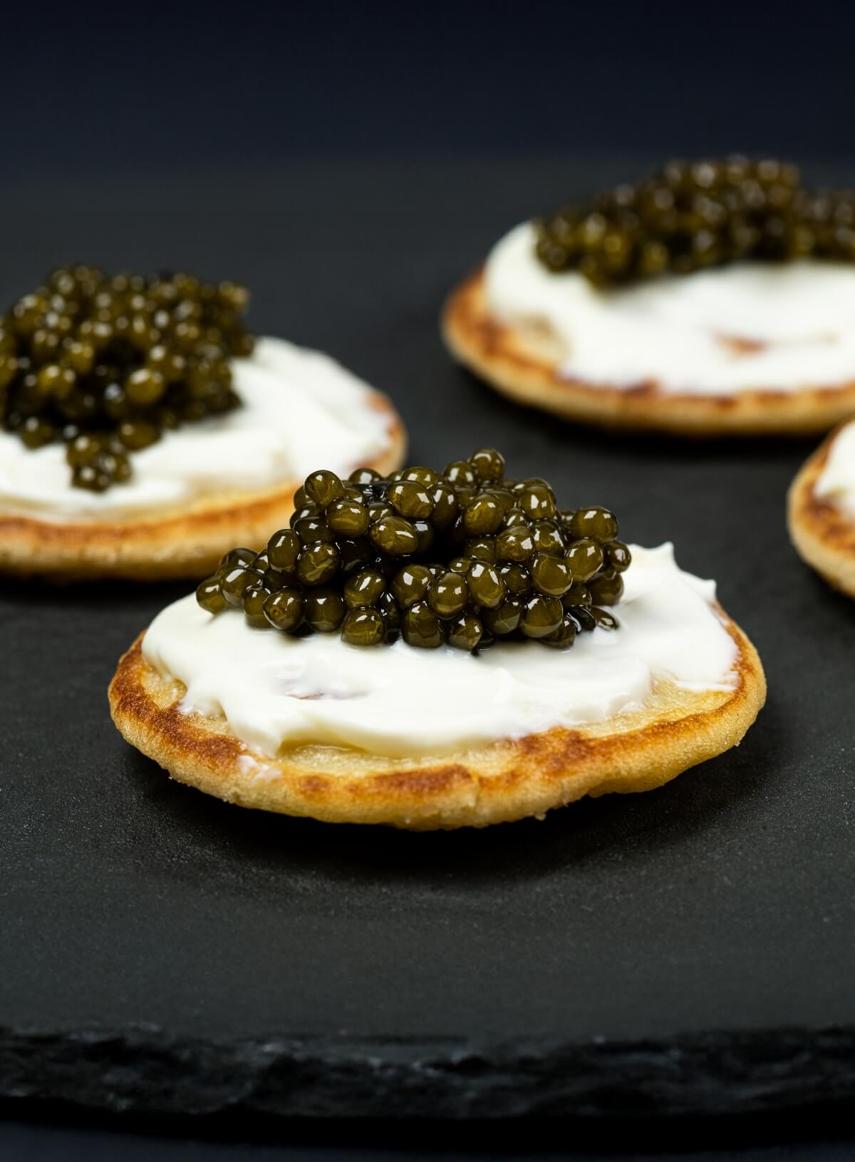 A close-up image of blini topped with luxury caviar, served on a dark surface.