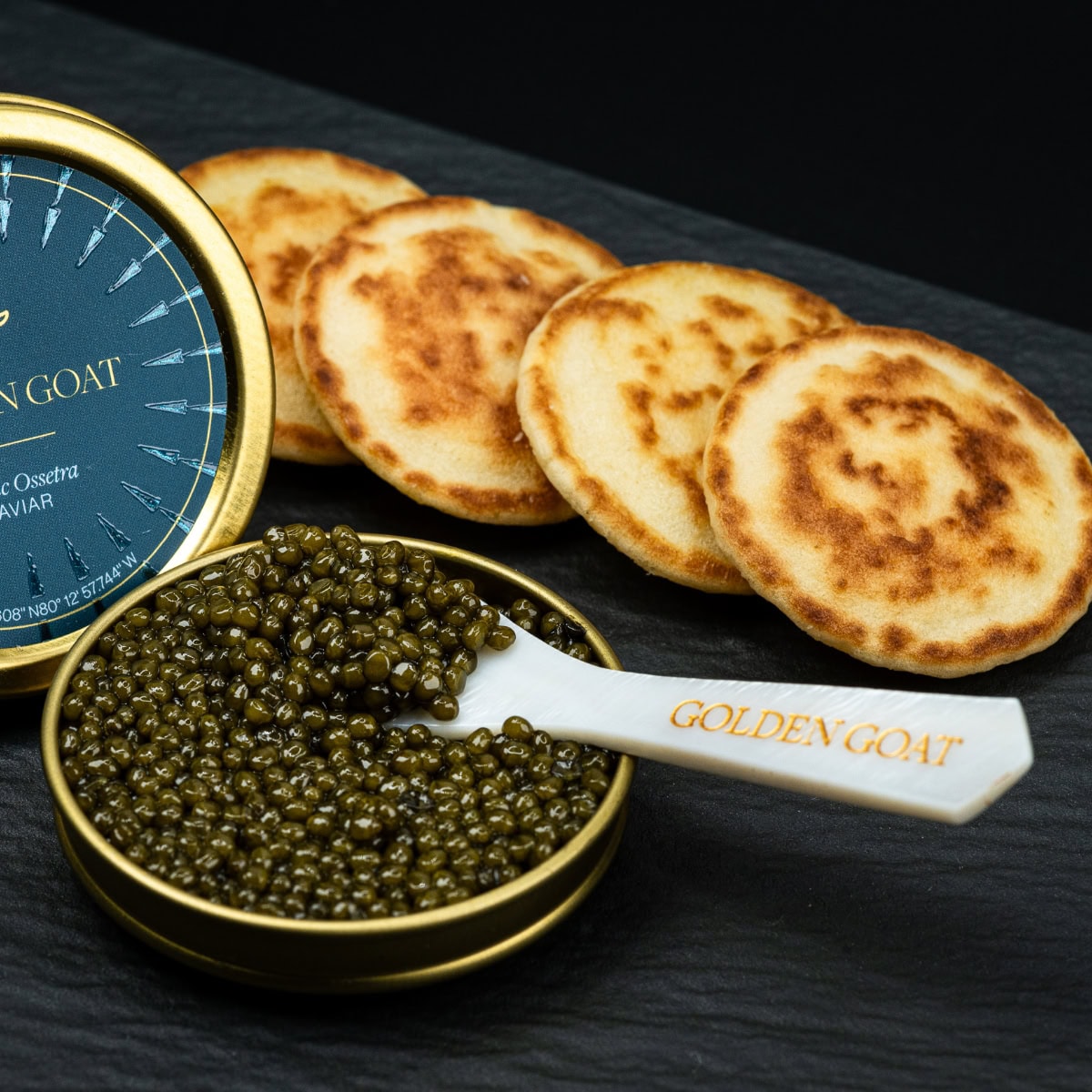 Tin of open caviar with a caviar spoon next to a row of blinis.
