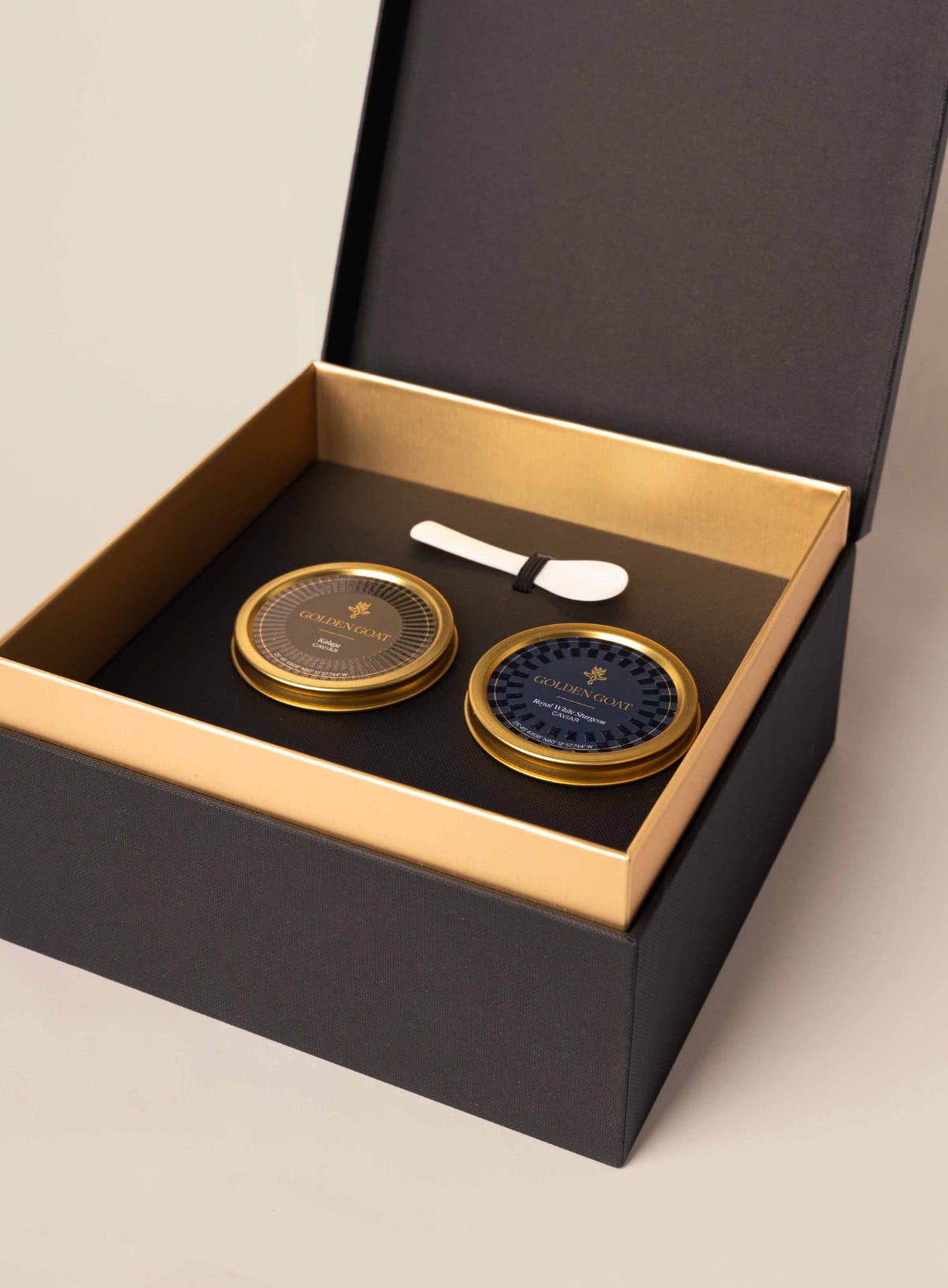 A luxurious golden goat caviar gift box open to reveal two tin containers of caviar, one labeled Kaluga and the other Royal White Sturgeon, accompanied by a small pearl spoon.