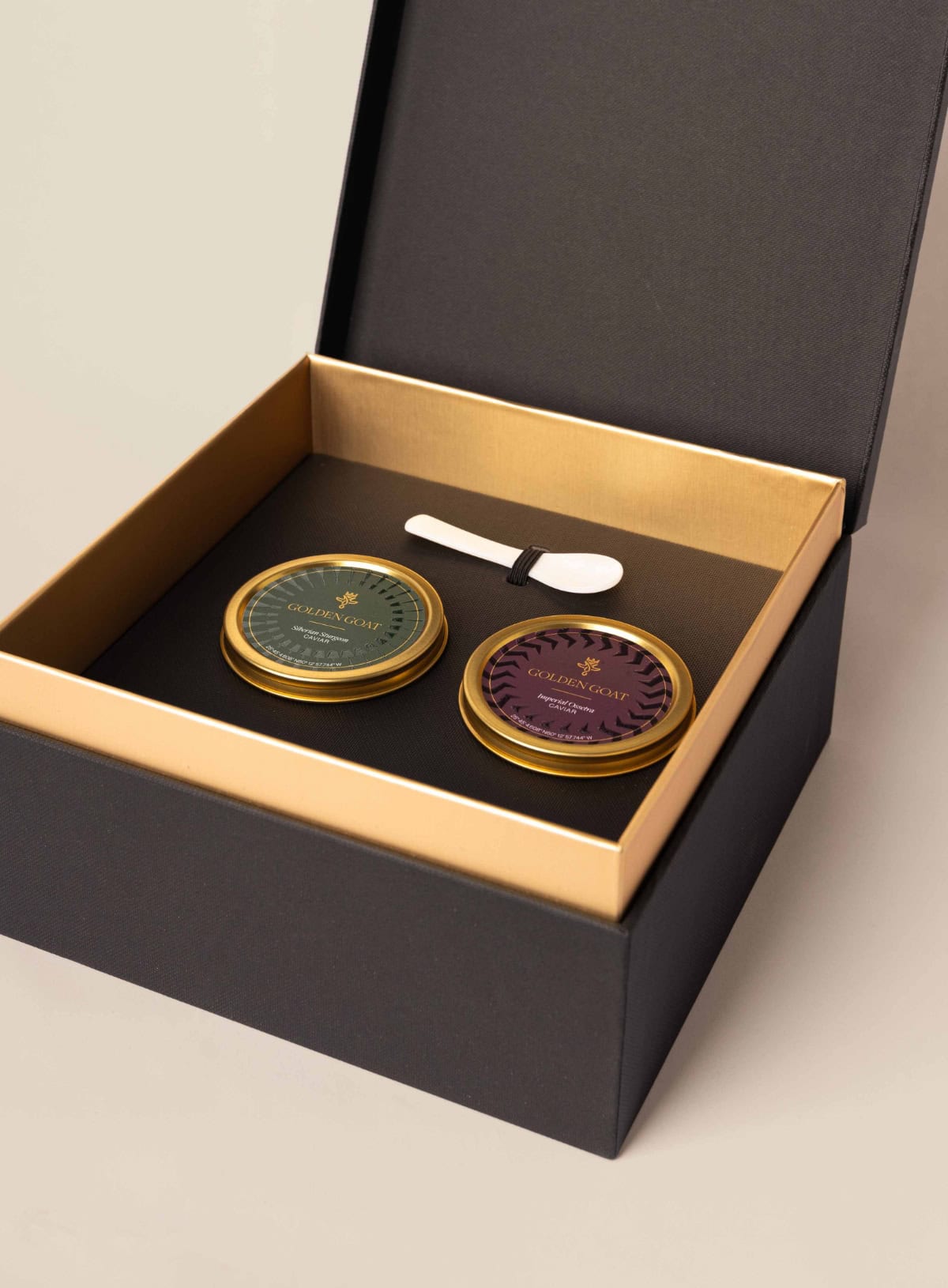 A luxurious golden goat caviar gift box open to reveal two caviar containers with a gold and purple label, alongside a small silver spoon.