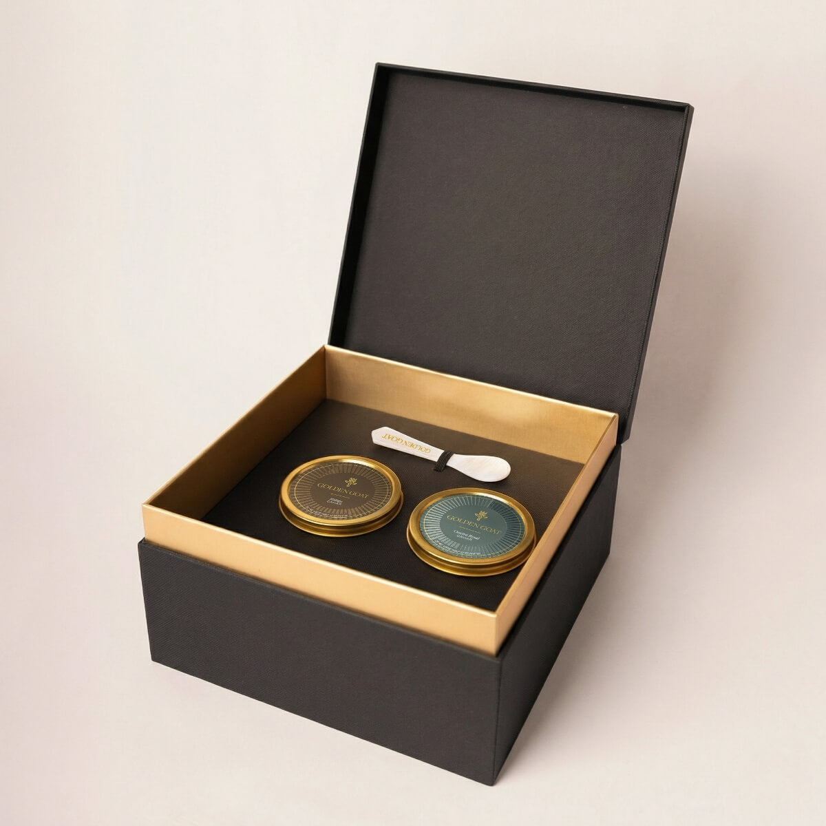 Two gold caviar tins with a white pearl spoon in a black and gold box on a light beige background