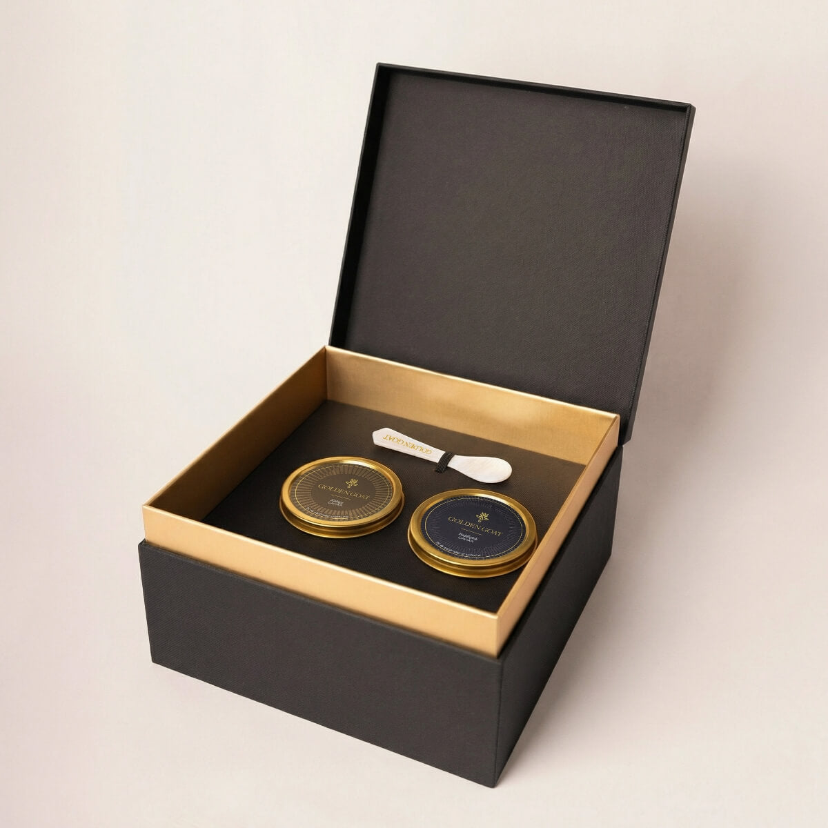 Two gold caviar tins with a white pearl spoon in a black and gold box on a light beige background