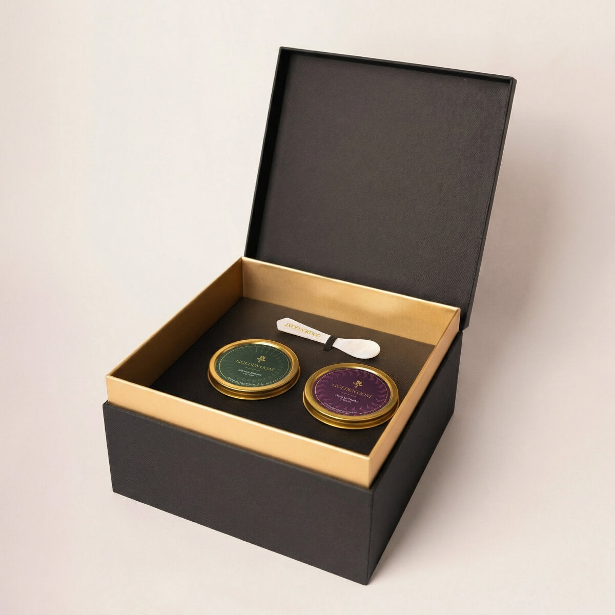 Two gold caviar tins with a white pearl spoon in a black and gold box on a light beige background