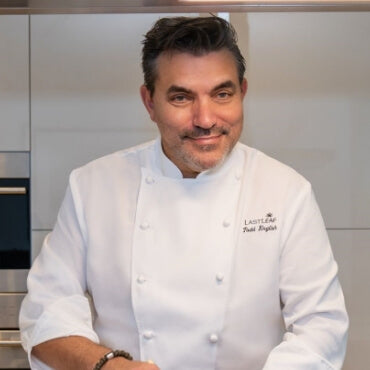 Chef in a professional kitchen wearing a white chef's coat with a visible brand logo.