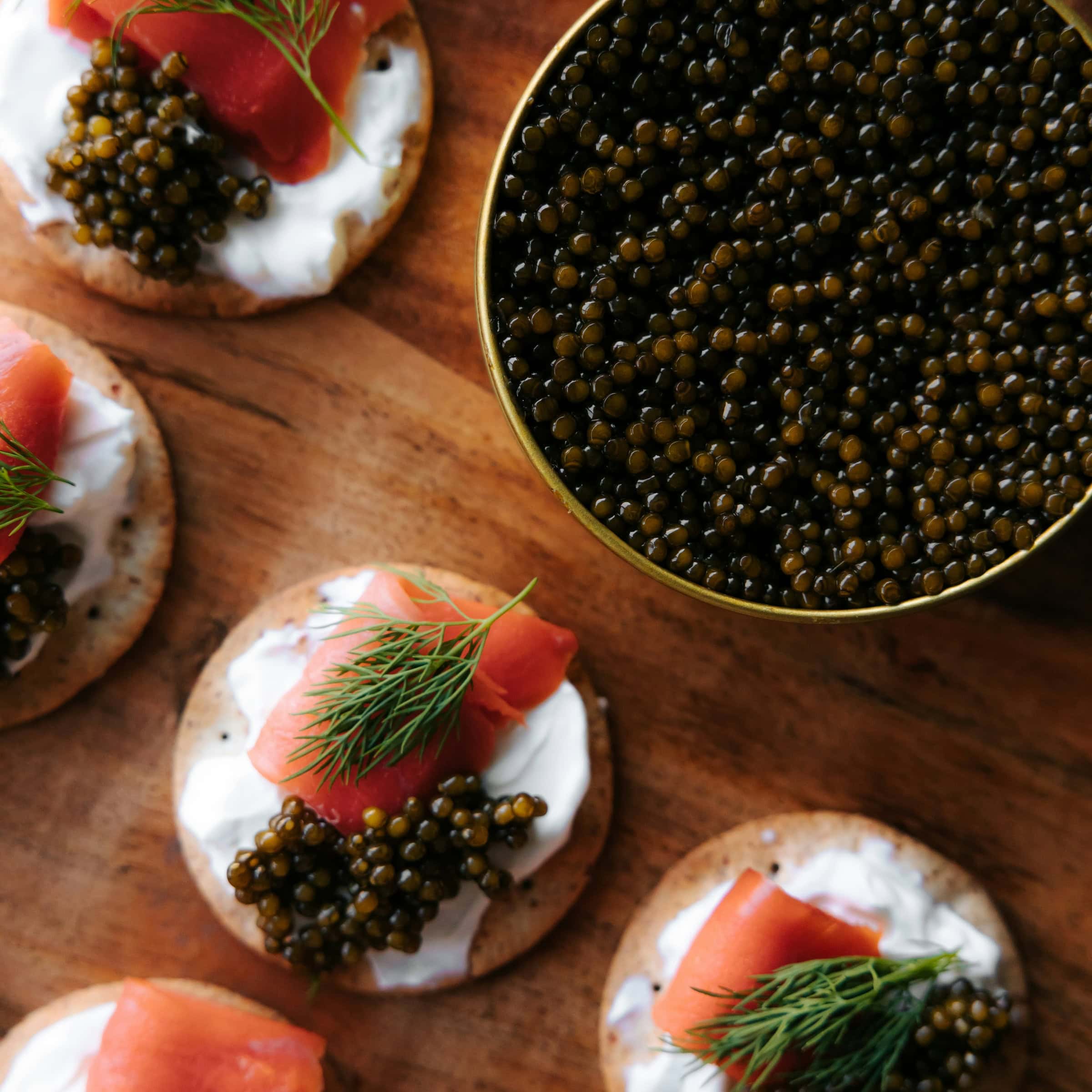 An open tin of caviar next to several crackers topped with cream, caviar and salmon on a wood table.