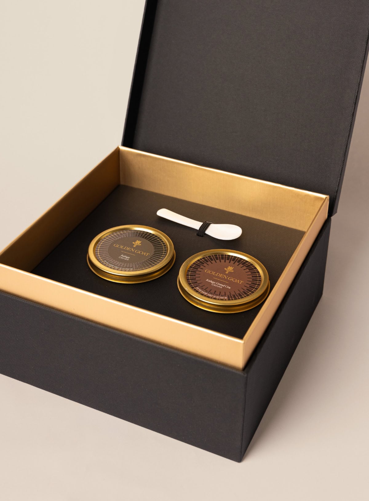 A luxurious gift box containing two jars of caviar, with a caviar key and pearl spoon placed on a golden tray inside the box.