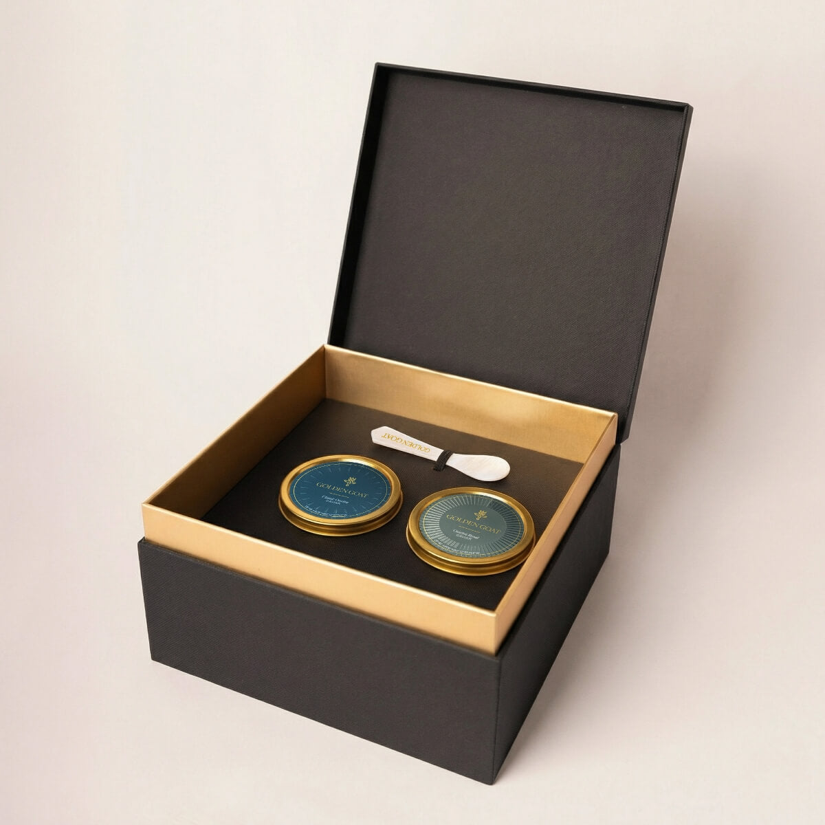 Two gold caviar tins with a white pearl spoon in a black and gold box on a light beige background