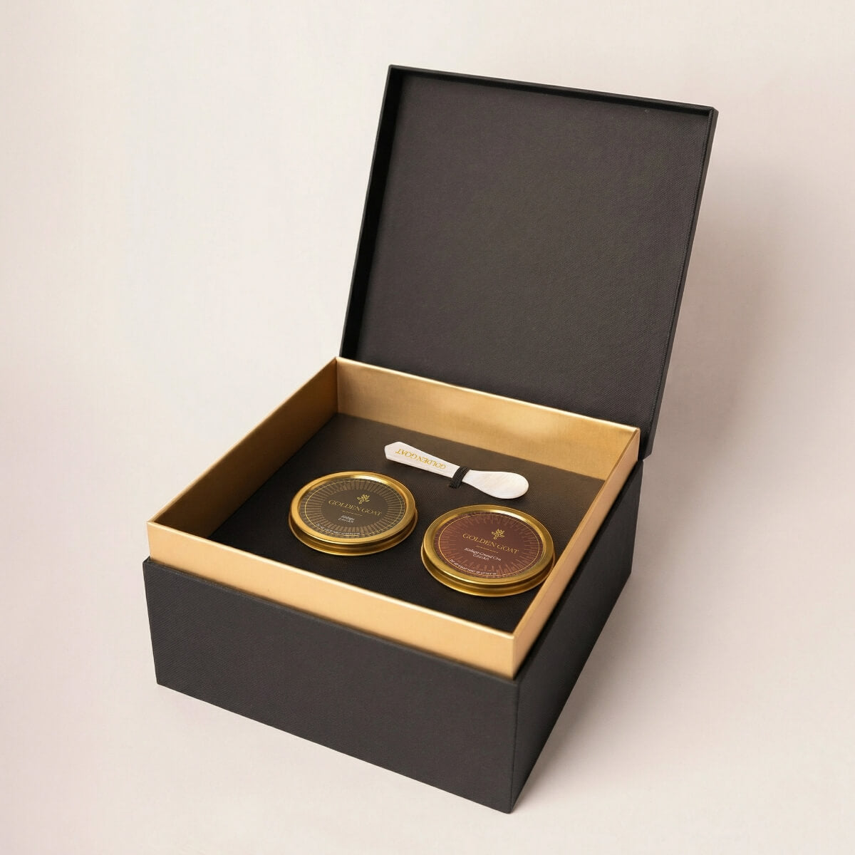Two gold caviar tins with a white pearl spoon in a black and gold box on a light beige background