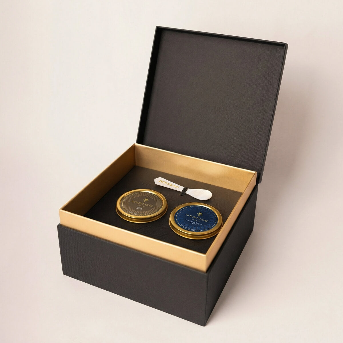 Two gold caviar tins with a white pearl spoon in a black and gold box on a light beige background