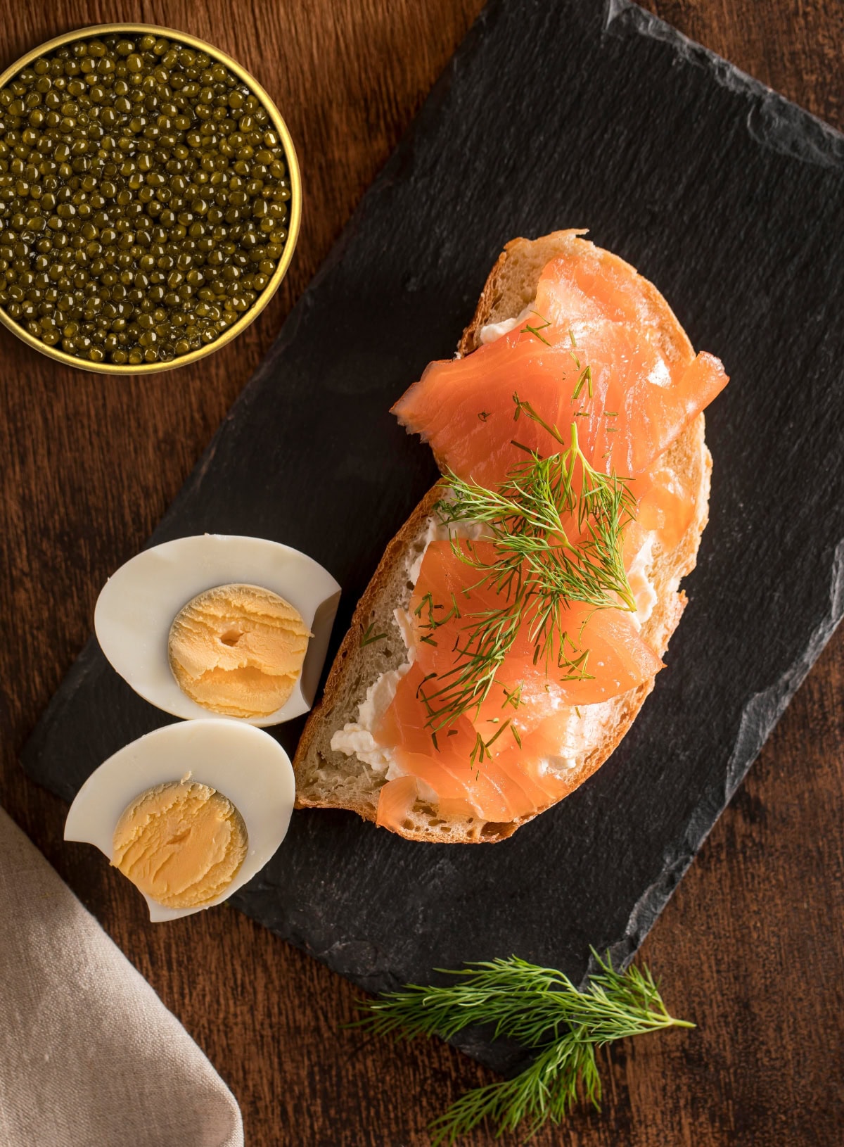 A slice of smoked salmon on a bagel, garnished with dill, alongside boiled egg slices and a dish of caviar in the background.