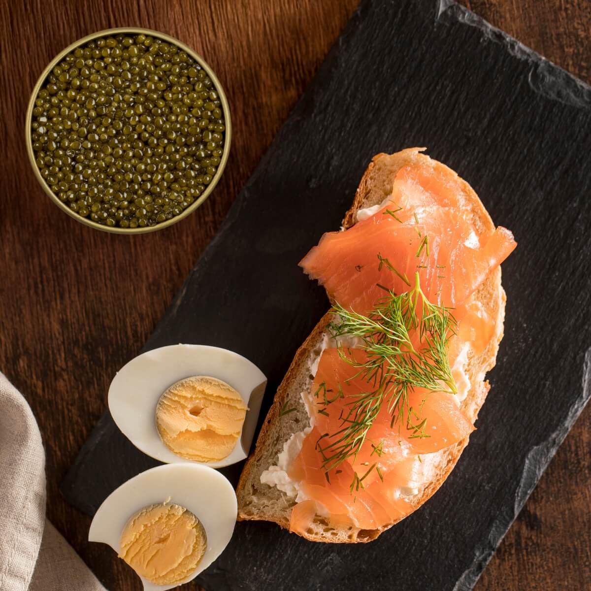 Smoked salmon on bread with cream cheese and dill, accompanied by a tin of caviar on a dark slate board.