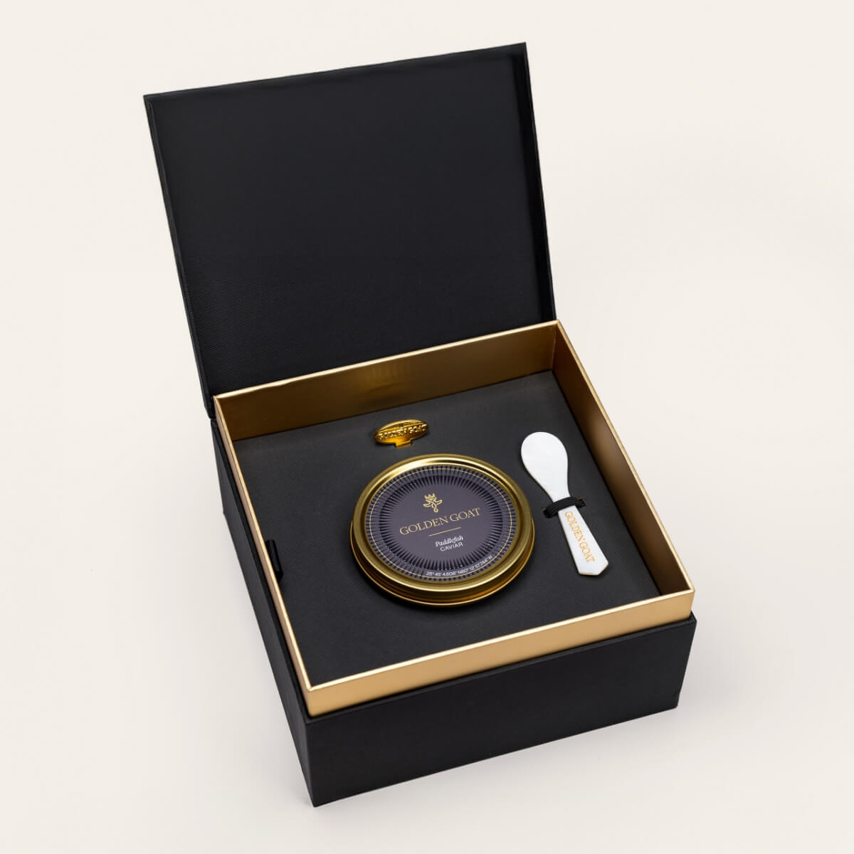 Black and gold box containing a gold caviar tin, caviar key, and pearl spoon on a white background.